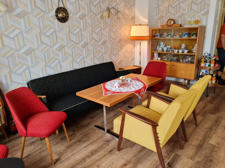 Sitzbereich im Cafe Miteinander in Geyer. Ein schwarzes Sofa, ein roter Stuhl und zwei gelbe Stühle. In der Mitte ein brauner Tisch. Im Hintergrund eine alte Komode mit Fenster von einer gemusterten Tapete