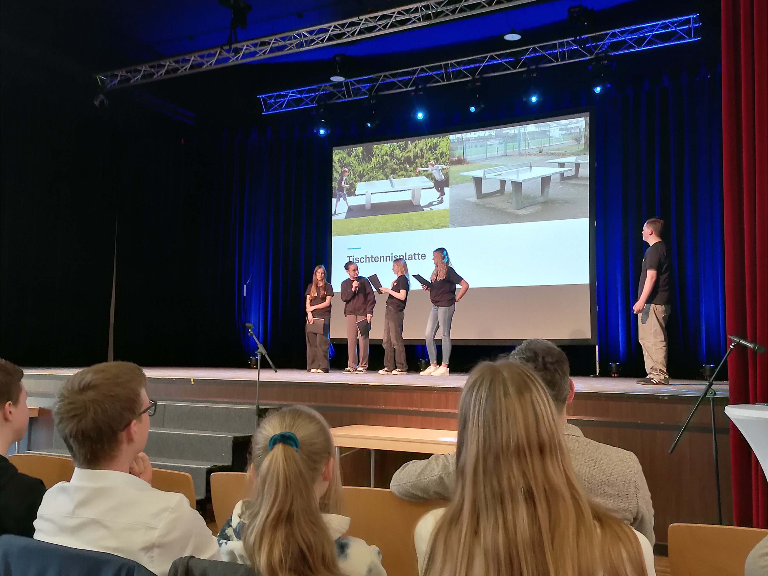 Auf dem Bild sieht man eine Bühne auf der fünf Personen stehen. Es sieht aus als wäre die rechte Person eine Art Moderator. Die weiteren vier Personen scheinen einen Vortrag zu halten. Hinter ihnen ist zudem eine Leinwand mit einem Bild zu sehen. Außerdem ist im Vordergrund des Bildes ein Teil des Publikums zu sehen. Auf dem Bild sieht man eine Bühne auf der fünf Personen stehen. Es sieht aus als wäre die rechte Person eine Art Moderator. Die weiteren vier Personen scheinen einen Vortrag zu halten. Hinter ihnen ist zudem eine Leinwand mit einem Bild zu sehen. Außerdem ist im Vordergrund des Bildes ein Teil des Publikums zu sehen.